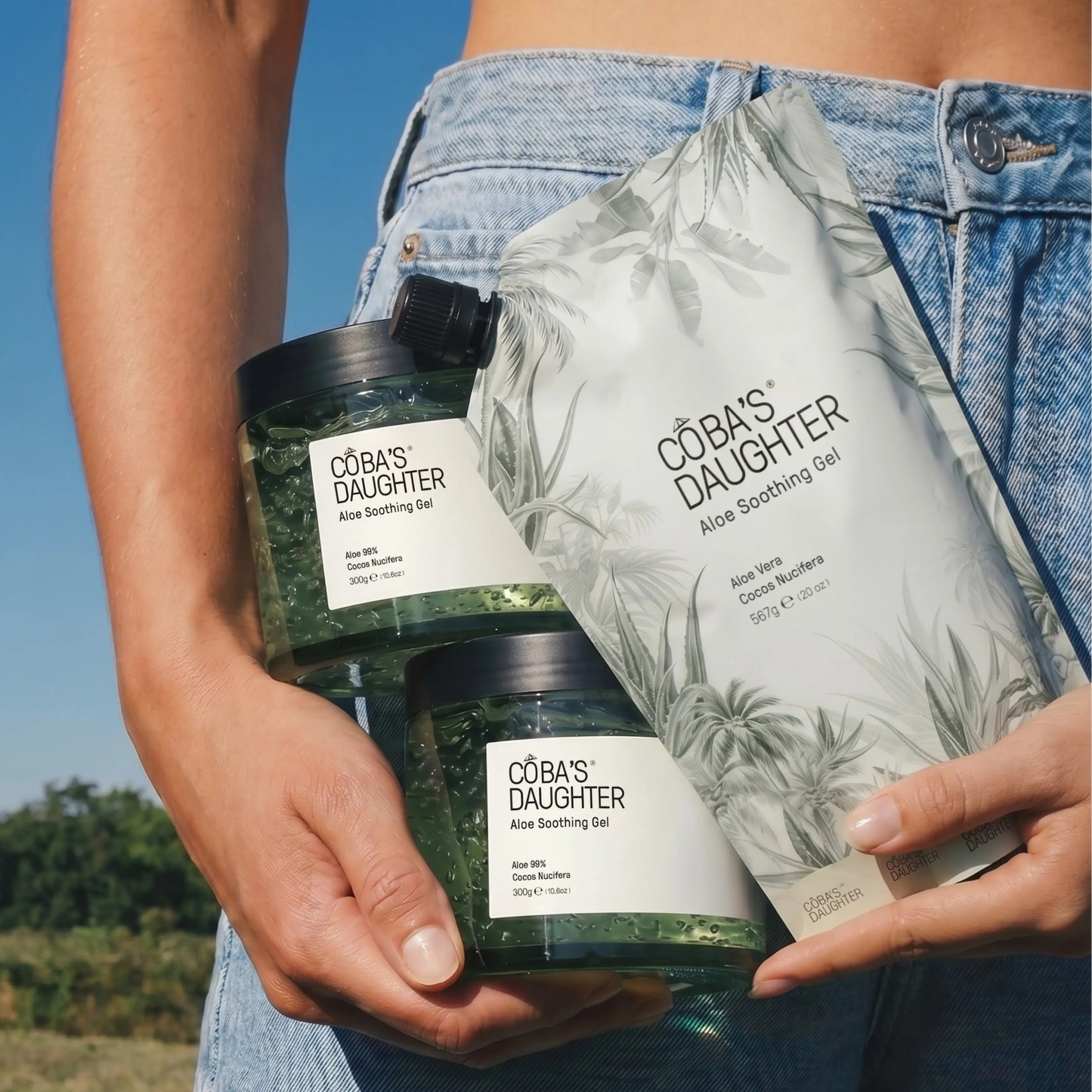 Woman wearing jeans holding CoBa's Daughter aloe gel jars and a refill pouch under the sun