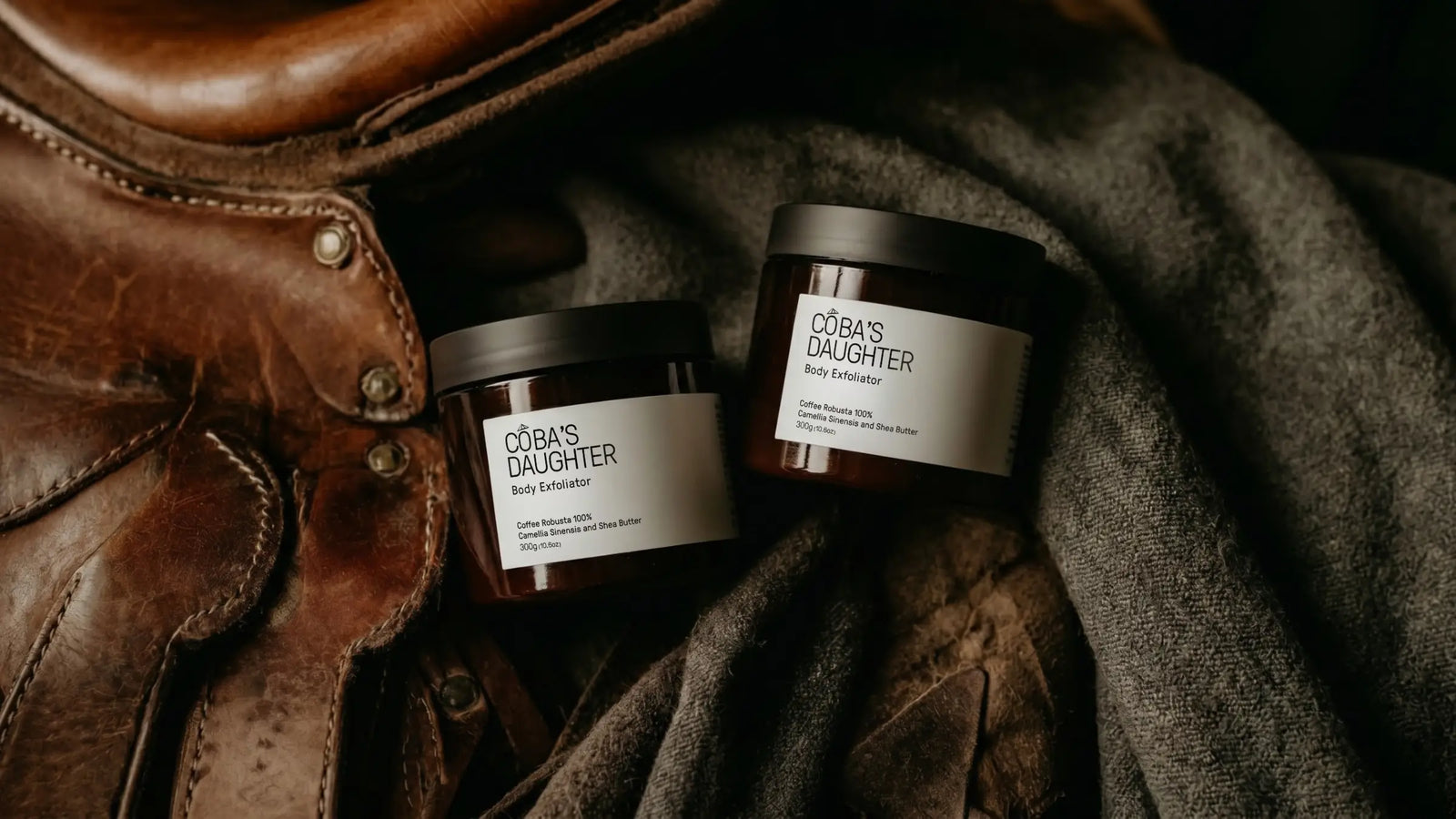 Two jars of CoBa's Daughter coffee body scrub on a textured surface with leather and fabric elements from equestrian, western style