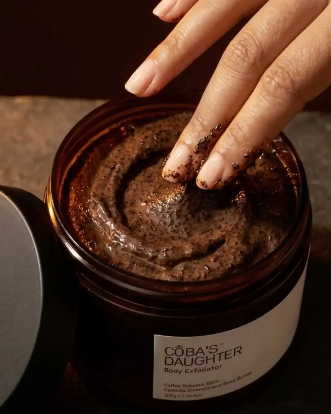 Jar of Coba's Daughter coffee body exfoliator with a hand touching the texture