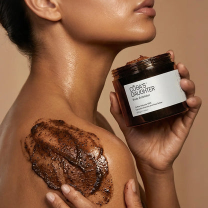 Applying CoBa's Daughter Robusta Coffee Body Scrub on shoulder showing dark brown coffee grounds texture for gentle exfoliation
