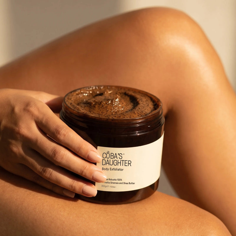 Applying CoBa's Daughter Robusta Coffee Body Scrub on shoulder showing dark brown coffee grounds texture for gentle exfoliation