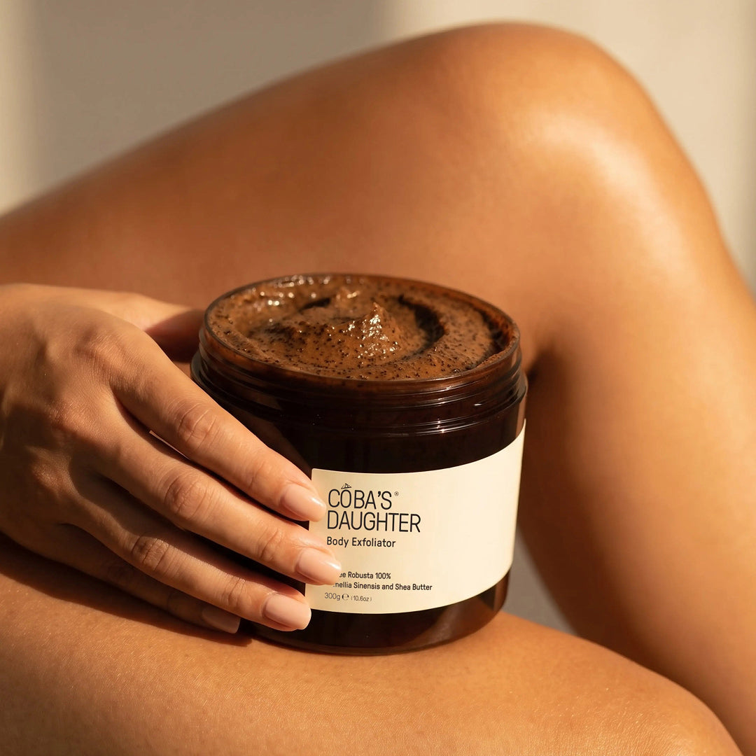 Hand holding an open jar of CoBa's Daughter Robusta Coffee Body Scrub against bare skin in warm light