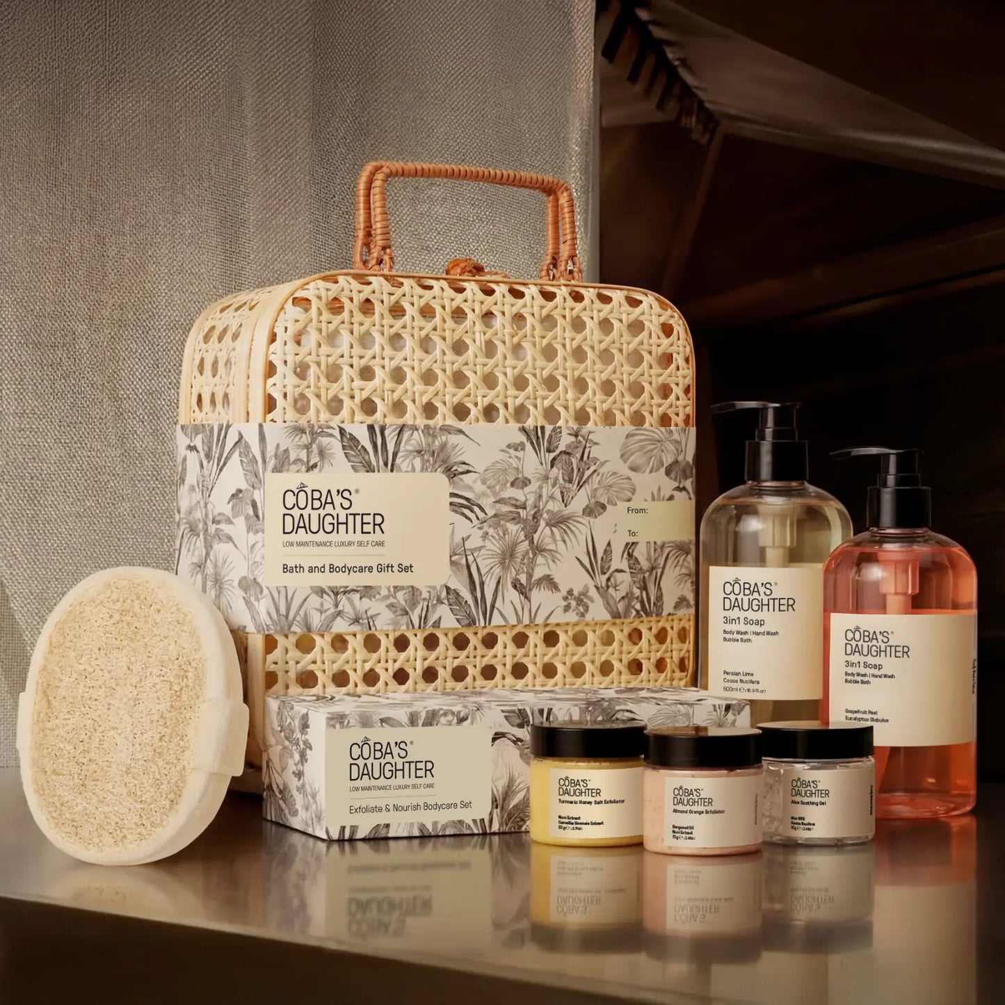 CoBa's Daughter bath and body products including a rattan basket, bottles, loofah sponge and on a reflective surface.