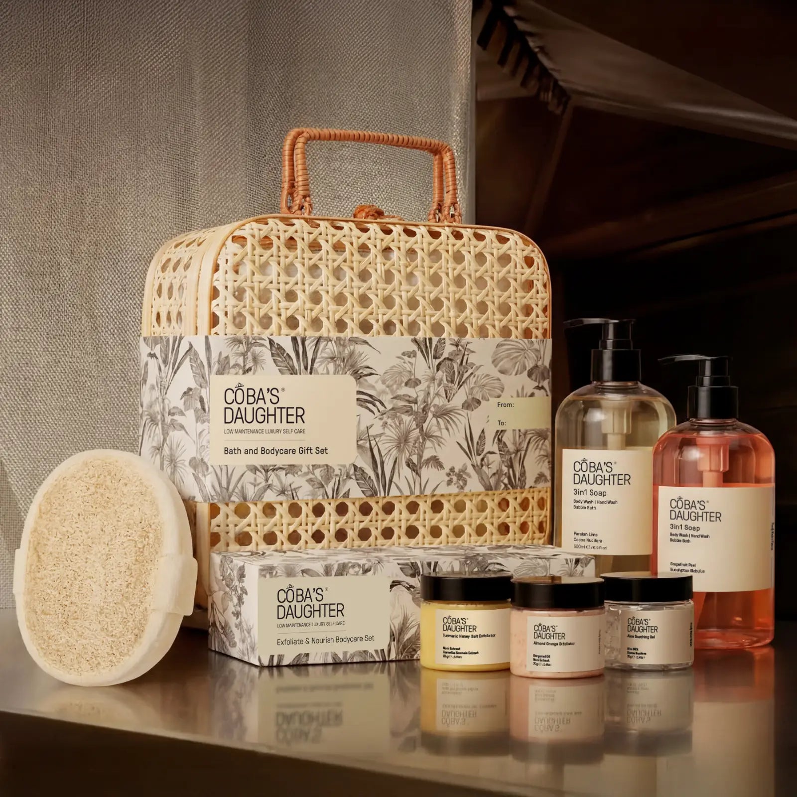 CoBa's Daughter bath and body products including a rattan basket, bottles, loofah sponge and on a reflective surface.
