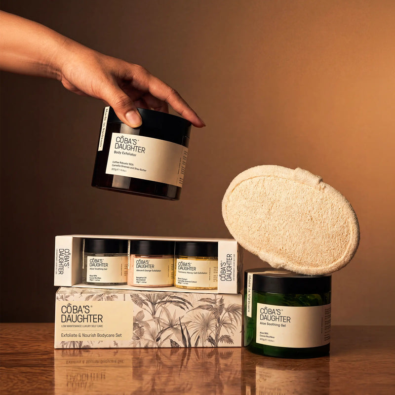 CoBa's Daughter body care products including coffee scrub, aloe gel, mini gift set, and a loofah on a wooden surface.