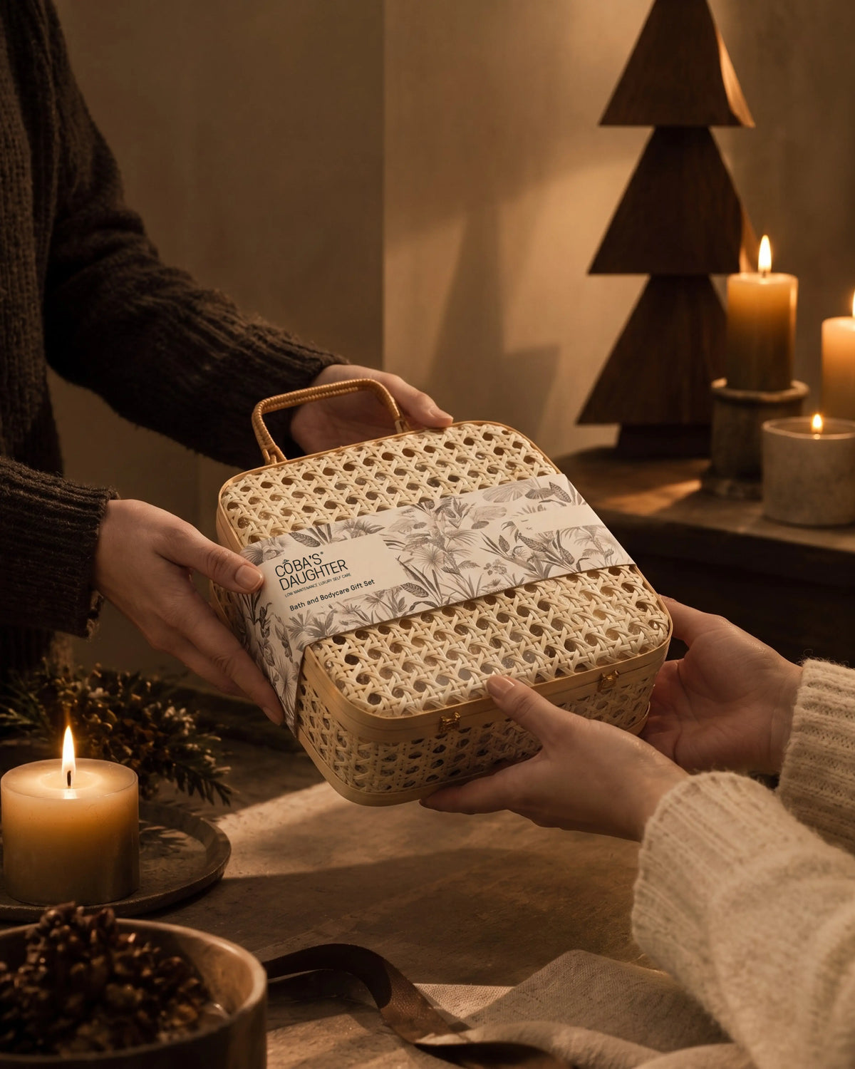 Two people passing a CoBa’s Daughter bath and body gift set in a rattan case across a candlelit table.