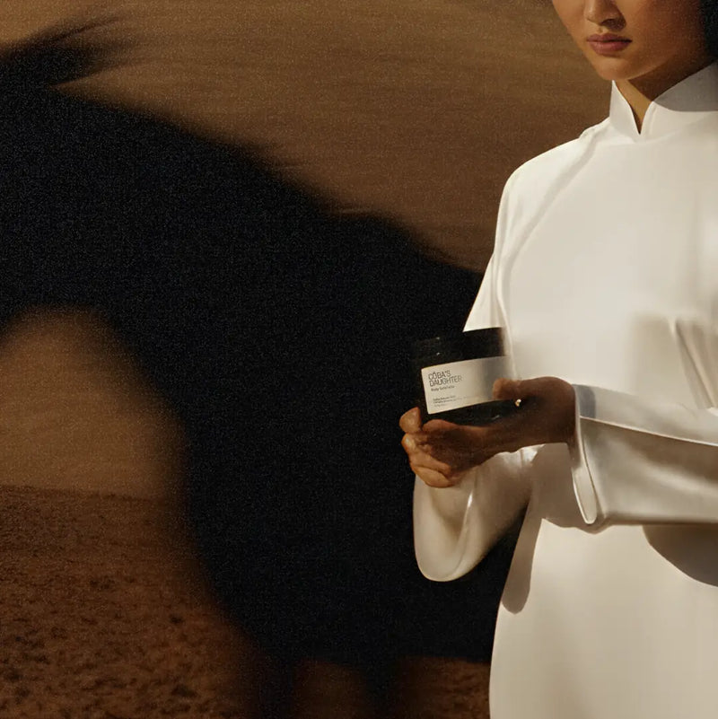 Model holding CoBa's Daughter Robusta Coffee Body Scrub jar in minimalist white outfit with horse in background dramatic shadow lighting
