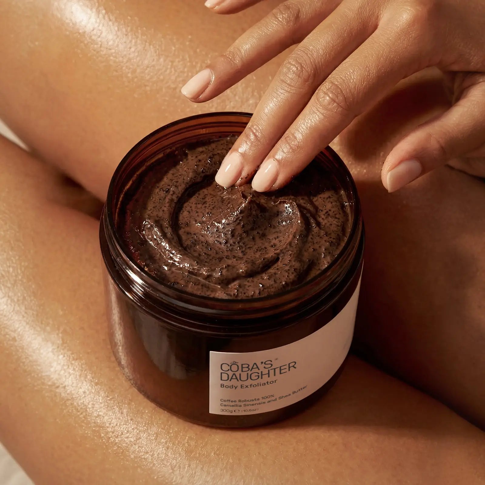 Scooping CoBa's Daughter Robusta Coffee Body Scrub from open jar showing dark brown finely milled texture with coffee grounds