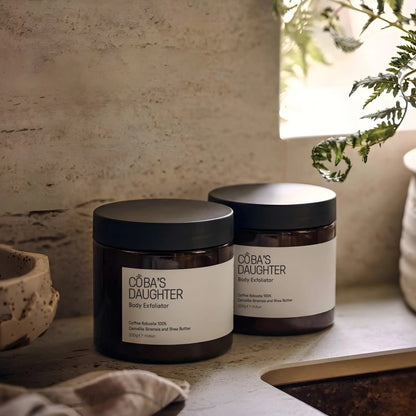 Two jars of CoBa's Daughter body exfoliator on a ceramic sink surface with a natural, ceramic background.