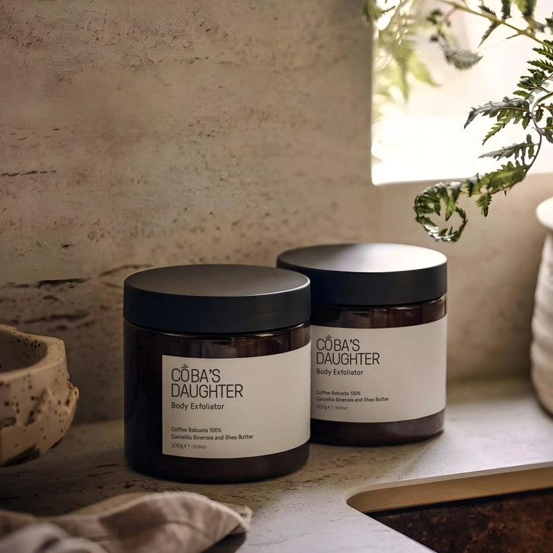 Two jars of CoBa's Daughter body exfoliator on a ceramic sink surface with a natural, ceramic background.